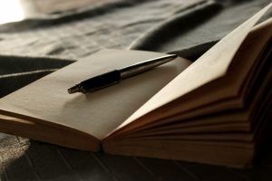 An open blank notebook rests on a dark surface, with a black-and-silver pen lying across its pages in warm, soft light.